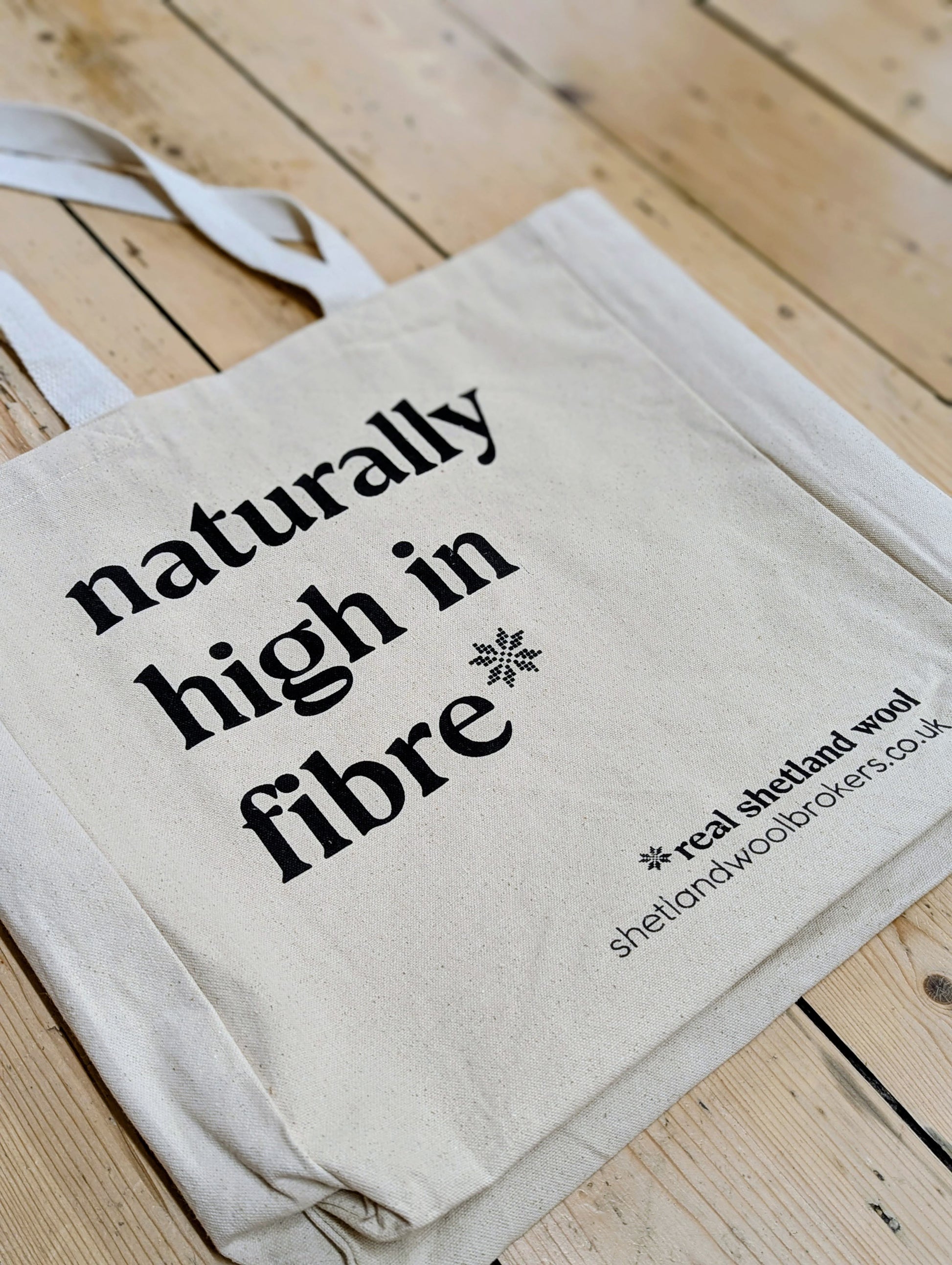 A natural canvas tote bag with black printed text reading 'naturally high in fibre' and a small snowflake graphic, placed on a light wood plank surface. The bag also includes the website text 'shetlandwoolsbrokers.